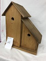 Photo of a rustic wooden bird house with two sections, each having a round entrance hole and pitched roofs, standing against a white backdrop.