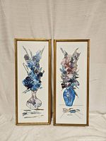 Two framed floral vase prints featuring blue and purple flowers in abstract vases, displayed upright against a curtain background.