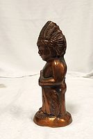 Side profile view of bronze Indigenous man figure with feather headdress, standing on rounded base.