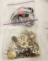 Two sealed zip-lock bags containing various costume jewelry pieces including faux pearls, metal chains, pendants, and bracelets.
