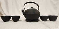 Full view of black cast iron Eastern style teapot placed on its stand, flanked by four matching teacups.