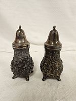 Front view of two intricately designed floral lead salt and pepper shakers, showing detailed raised floral patterns and metal tops.