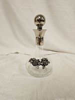 Clear glass decanter bottle with silver collar and round stopper, decorated with metal grape clusters at base, photographed on white fabric background.