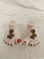 View of the front and top of two ceramic foot-shaped salt and pepper shakers with pineapple Hawaii decal and pink toenails.