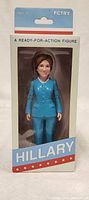 Front view of boxed Hillary Clinton action figure showing the figure in blue suit and product name 'HILLARY' on packaging.
