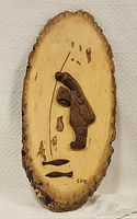 Wooden decorative plaque depicting Indigenous fisherman with fishing rod and two fish, mounted on natural wood slice with bark edges, signed 'L.A.R.'