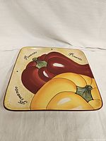 Front view of square ceramic plate with painted red and yellow pimento peppers and 'Pimento' text, beige background with brown border.