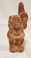 Frontal view of a small reddish-orange and beige clay figure with stylized human features, an ornate headdress, and carved design details.