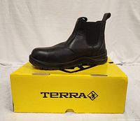 Side view of single black Terra boot placed on original yellow Terra box, showing leather upper, elastic side panel, rugged sole and pull tab on back.