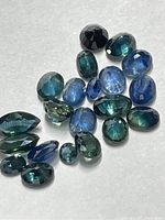 Photo of 22 loose faceted sapphire gemstones in blue and greenish-blue hues on white background.