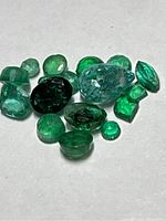 Close-up photo of a collection of 15 loose natural emerald gemstones varying in shape, size, and shade of green.