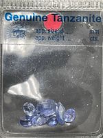Photo showing multiple small tanzanite gemstones in a labeled plastic packet indicating 'Genuine Tanzanite' with approximate weights and sizes not filled in.