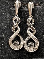 Front view of two intertwined loop sterling silver earrings with black and white diamonds visible on the curved surfaces and a black diamond cluster near the bottom.