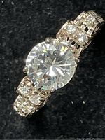 Close-up image showing large round clear cubic zirconia center stone with smaller CZ stones on silver band against black background.