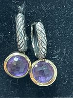 Close-up image of two sterling silver earrings with faceted round purple amethyst stones and twisted rope design on silver hoops. Earrings are shown resting on black fabric background.
