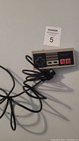 Photo of an original Nintendo Entertainment System wired controller with attached black cable and proprietary NES connector plug against a gray background.