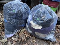 Two large clear plastic bags filled with assorted used men’s and women’s clothing placed outside on dry leaves.