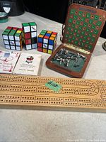 Photo showing 3 Rubik's cubes, wooden cribbage board with pegs, 3 decks of cards and small chess board with pieces.