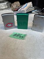 Front view of three Zippo lighters; one green with cannabis leaf, one silver with Montreal Canadiens logo, one plain silver.