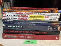 Stack of 11 books including Warhammer 40,000 titles, manga drawing guides, and Palladium role-playing game book taken from front showing book spines and titles.
