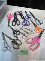 14 pairs of scissors in various sizes, colors, and handle shapes laid out showing condition and variety.