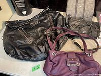 Three purses shown together: large black Guess purse with braided handles, smaller black Nine West purse, and purple Rosetti purse.