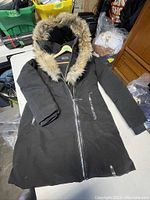 Black women's long winter coat laid flat on table, showing full coat with zipped front and faux fur hood trim.