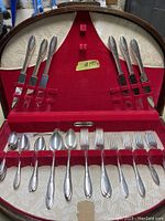 Opened wooden box displaying antique silver plated knives, forks, and spoons held in red velvet slots with white fabric lining inside lid.