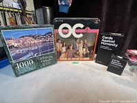 Three items on a table: a 1000-piece puzzle box with Côte d'Azur marina scene, The OC game box with TV show characters, and a Cards Against Humanity game set.