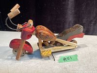 Side angle photo showing the handmade wooden motorcycle sculpture with red front fender, detailed woodburned fuel tank, black seat, wooden wheels, and handlebars with black wiring.