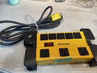 Top view of yellow Mastercraft power bar showing seven power outlets, red power switch, and attached long power cord with yellow plug on a grey surface