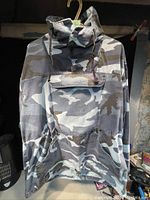 Front view of men's camouflage hoodie hanging on hanger showing pullover style, front pouch pocket with flap, drawstring collar, and camouflage pattern in grey tones.