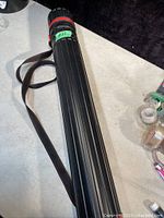 Black expandable poster storage tube lying on a surface, featuring red locking cap and attached brown adjustable shoulder strap.