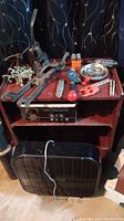 Brown wooden cabinet with tools arranged on top shelf including clamps, cutters, screwdriver, metal bowls, and light bulbs. Middle shelf has a black metal control box. Below is a large black box fan with electrical cord.