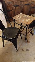 Side view of black metal chair next to vintage rolling cart with worn wooden top and metal frame with casters.
