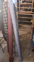 Photo showing four rolls of fabric in different colors and patterns leaning against a vintage wooden drying rack in a storage or workshop area.