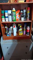 Brown wooden shelf with various spray cans and chemicals on top and middle shelves, atop a light-colored cabinet with drawers below.