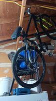 Photo of black mountain bicycle hanging from ceiling hooks in garage. Visible details include yellow handlebar grips and knobby tires.