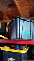 Large black plastic storage tote container on a shelf with lid closed, blue label attached