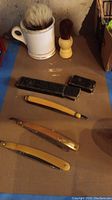 Photo showing three vintage straight razors with yellow and amber handles, two black storage cases, and two shaving brushes with light and dark bristles.