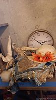 Photo showing a portion of the wall clock, porcelain bird and human figurine, decorative pillow with tassels, and white straw hat with orange flowers.