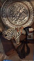 Photo showing metal decorative plate with embossed battle scene, wooden zebra figurine, cast iron trivets, and perfume bottles.