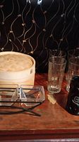 Photo showing bamboo steamer with lid, three tall drinking glasses and part of the glass condiment holder on a wooden surface.
