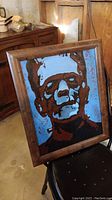 Full view of framed abstract painting of Frankenstein's Monster with brown wooden frame, set against background of wooden furniture and concrete floor.