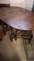 Photo showing the oval wooden antique gateleg table with two fold-down ends, turned legs connected by stretchers, surface shows wear.