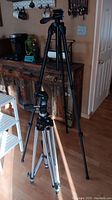 Two large tripods standing on the floor, one shorter with silver legs and one taller with black legs.