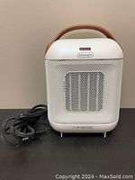 Photo of DeLonghi Capsule heater front view showing white plastic body, mesh grille, power cord coiled beside the unit, and carrying handle on top.