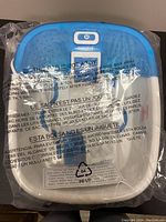 Top view of the foot spa in blue and white, showing power button and raised nodes, wrapped in a plastic bag with safety warnings in multiple languages.