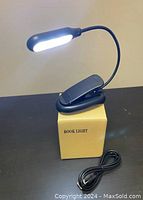 Black portable LED book light attached to cardboard box with USB cable coiled beside it, shown lit up with white LEDs.