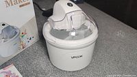 White VPCOK ice cream maker with clear lid and control button on top, shown on kitchen counter with box.
